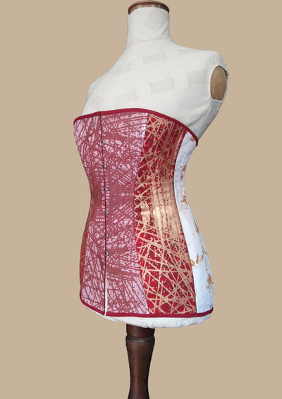 Corset. Front/side view. White cotton dyed red and red fine wool screen printed with cross linear pattern in copper and red tones, white cotton rust dyed, red cotton drill, steel split busk, steel spiral boning and rigilene boning.