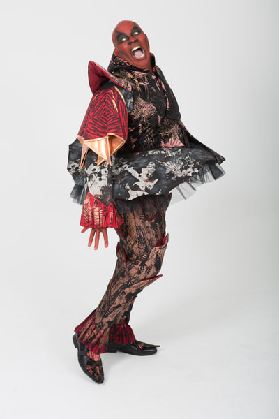 Costume for Mephistopheles in Faust. Handmade. Slashed trousers, sleeveless doublet with shaped collar, cote with flared skirt and bum roll, pleated cuffs on sleeves and ankle. Tones of red, black, gold and copper with white bleach effect patterning. Front  view. Performer: G. Stewart (male).