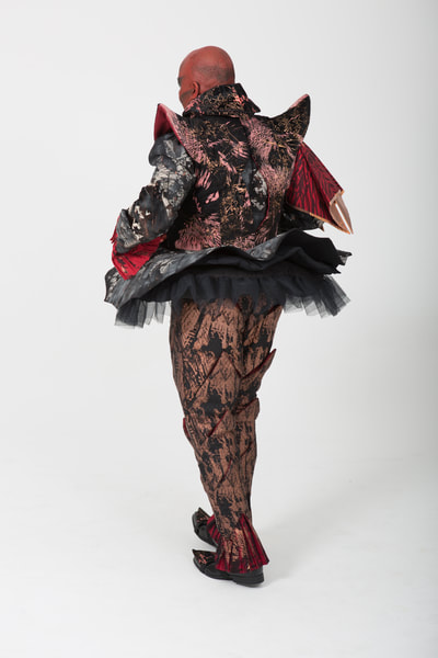 Costume for Mephistopheles in Faust. Handmade. Slashed trousers, sleeveless doublet with shaped collar, cote with flared skirt and bum roll, pleated cuffs on sleeves and ankle. Tones of red, black, gold and copper with white bleach effect patterning. Back  view. Performer: G. Stewart (male).